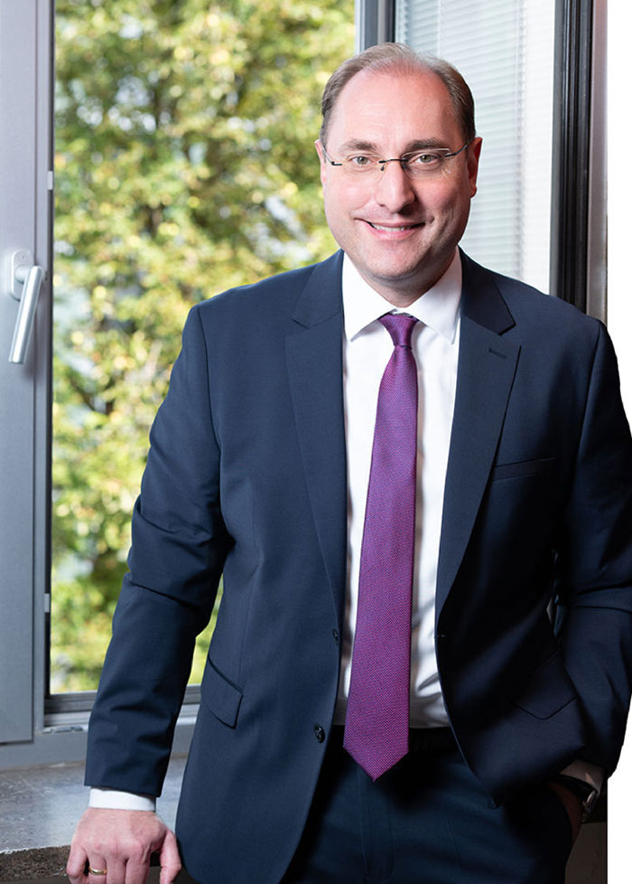Porträt Steuerberater Hoffmann im Anzug im Außenbereich vor unscharfem Hintergrund.